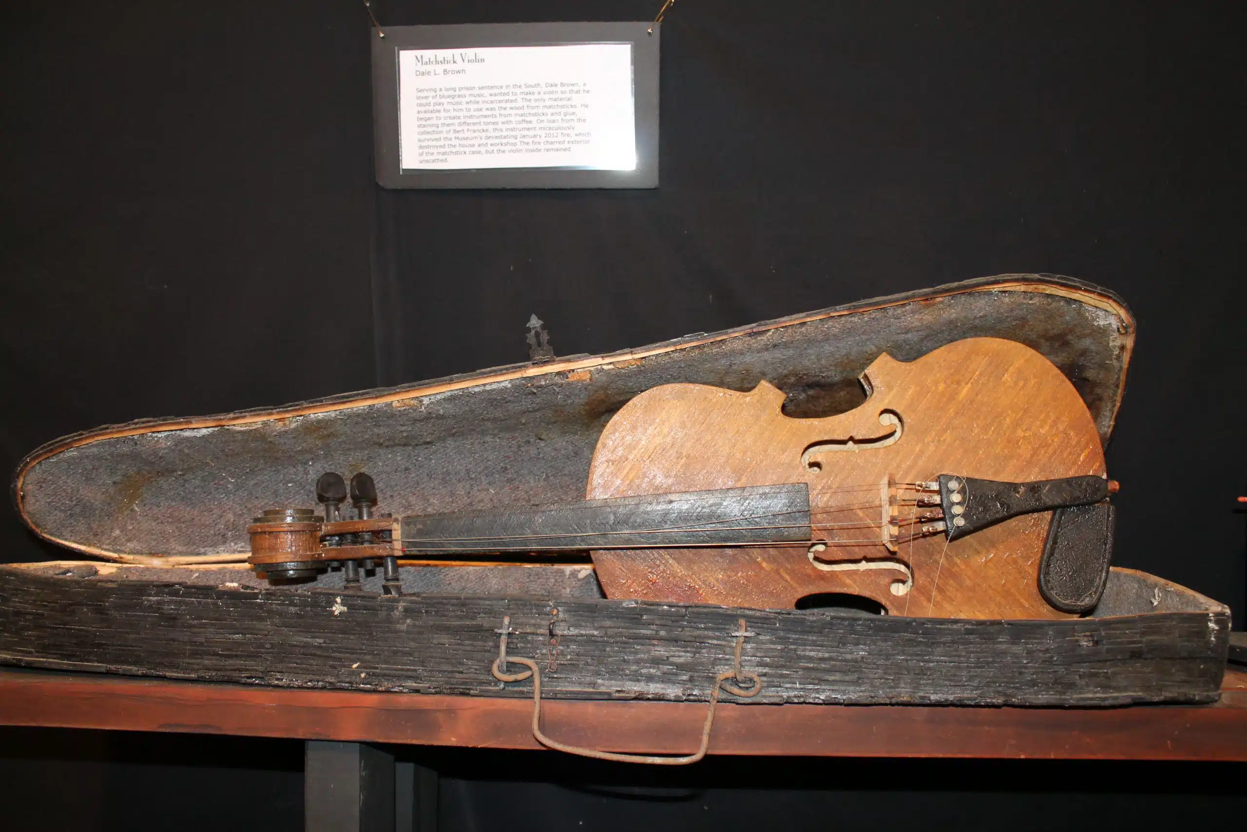 A weathered violin rests in an aged, partially open case on a dark wooden surface with an informational plaque in the background.