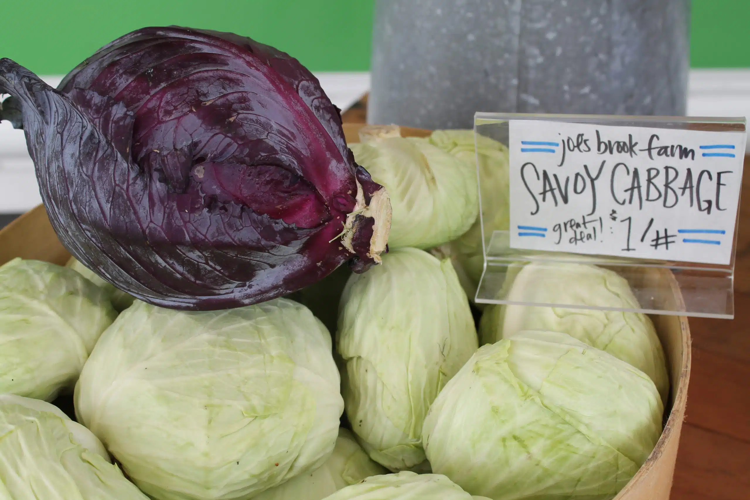 A basket filled with green and purple cabbages is displayed at a market, with a sign reading "Savoy Cabbage, 1$/lb.
