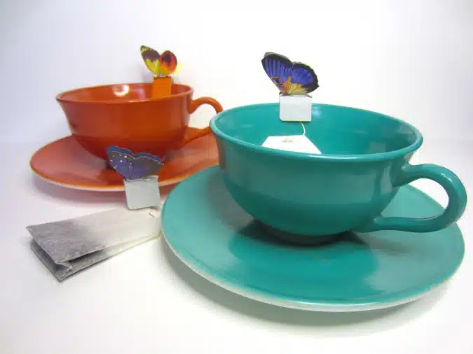 Two teacups with saucers, one orange and one teal. Each cup contains a teabag with a butterfly-shaped tag. Two unused teabags are placed next to the cups. White background.