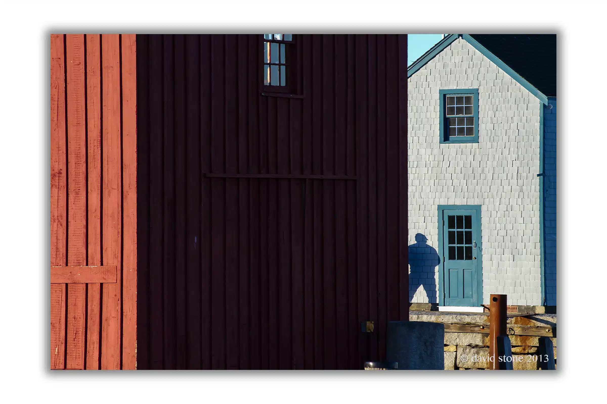 A person’s shadow is cast on a grey building with a blue door, partially hidden by a large red wooden structure in the foreground. The image is credited to David Stone, 2013.