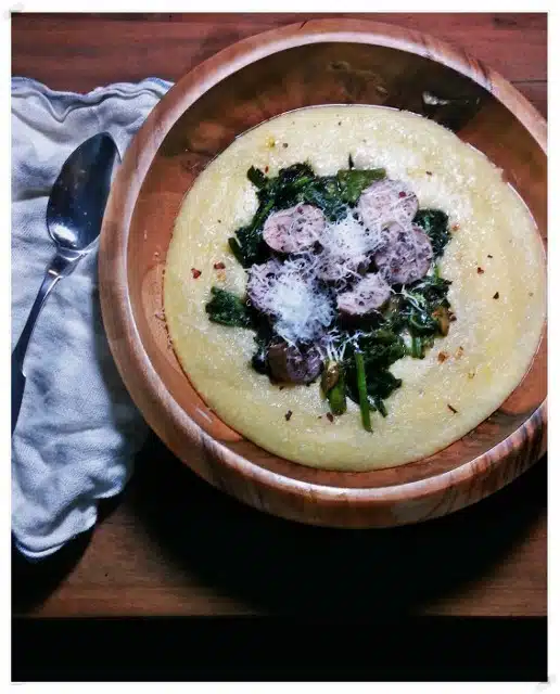 A wooden bowl filled with polenta, topped with sautéed greens and sliced sausage, sprinkled with grated cheese. A cloth napkin and a spoon are beside the bowl.