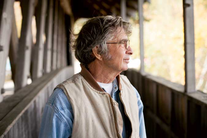 An older man with glasses and grey hair stands on a covered wooden bridge, looking to the side. He is wearing a beige vest over a blue shirt.