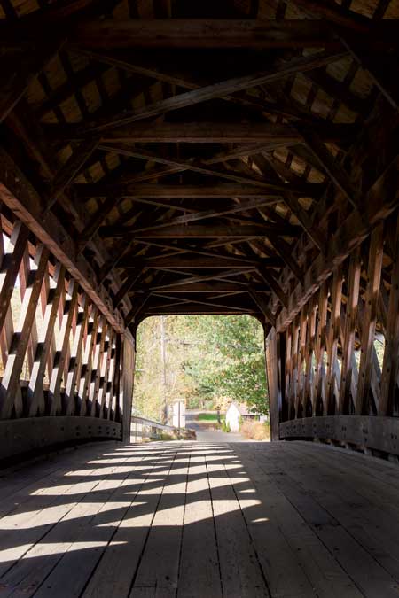 A wooden covered bridge with diagonal lattice sides and a plank floor. Sunlight filters through the openings, casting shadows on the walkway. Trees can be seen at the far end of the bridge.