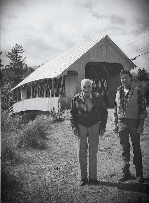 An older and younger man stand in front of a covered bridge in a rural setting.