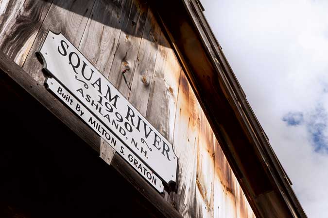A wooden sign reads "Squam River, 1990, Ashland N.H., Built by Milton S. Graton" on the side of a weathered wooden structure under a partly cloudy sky.