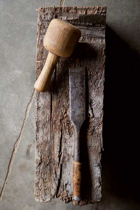 A wooden mallet and a chisel lie on a weathered piece of wood on a concrete surface.