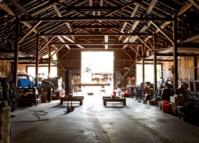Interior of a spacious, well-lit barn with various tools, equipment, and vehicles. The open barn doors offer a glimpse of the outside area on a sunny day.