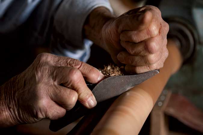 Close-up of an older person’s hands using a woodworking tool to carve a piece of wood, with shavings being removed from the surface.