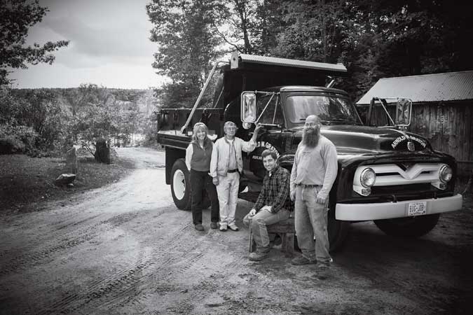 Four people stand and sit around an old dump truck on a dirt path with trees and a wooden building in the background.