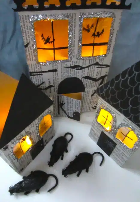 Three paper houses decorated with bat silhouettes and illuminated from inside with orange light; three black plastic rats are placed in front.