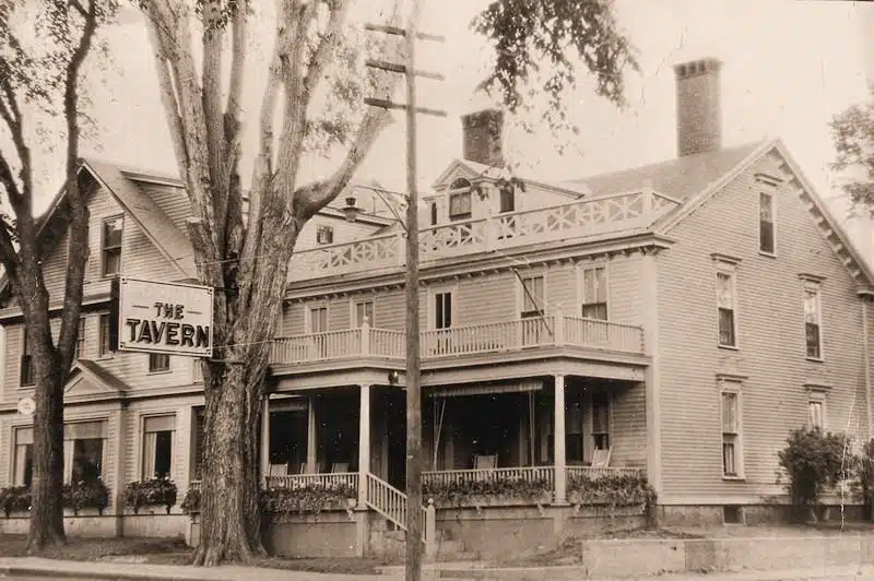 Sepia-toned photograph of a large, two-story wooden building with a sign reading "THE TAVERN" hanging from a pole in front. The building features multiple balconies and a porch, with trees nearby.