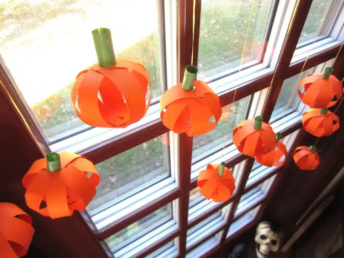 Orange paper pumpkins hang from strings in front of a window with a view outside.