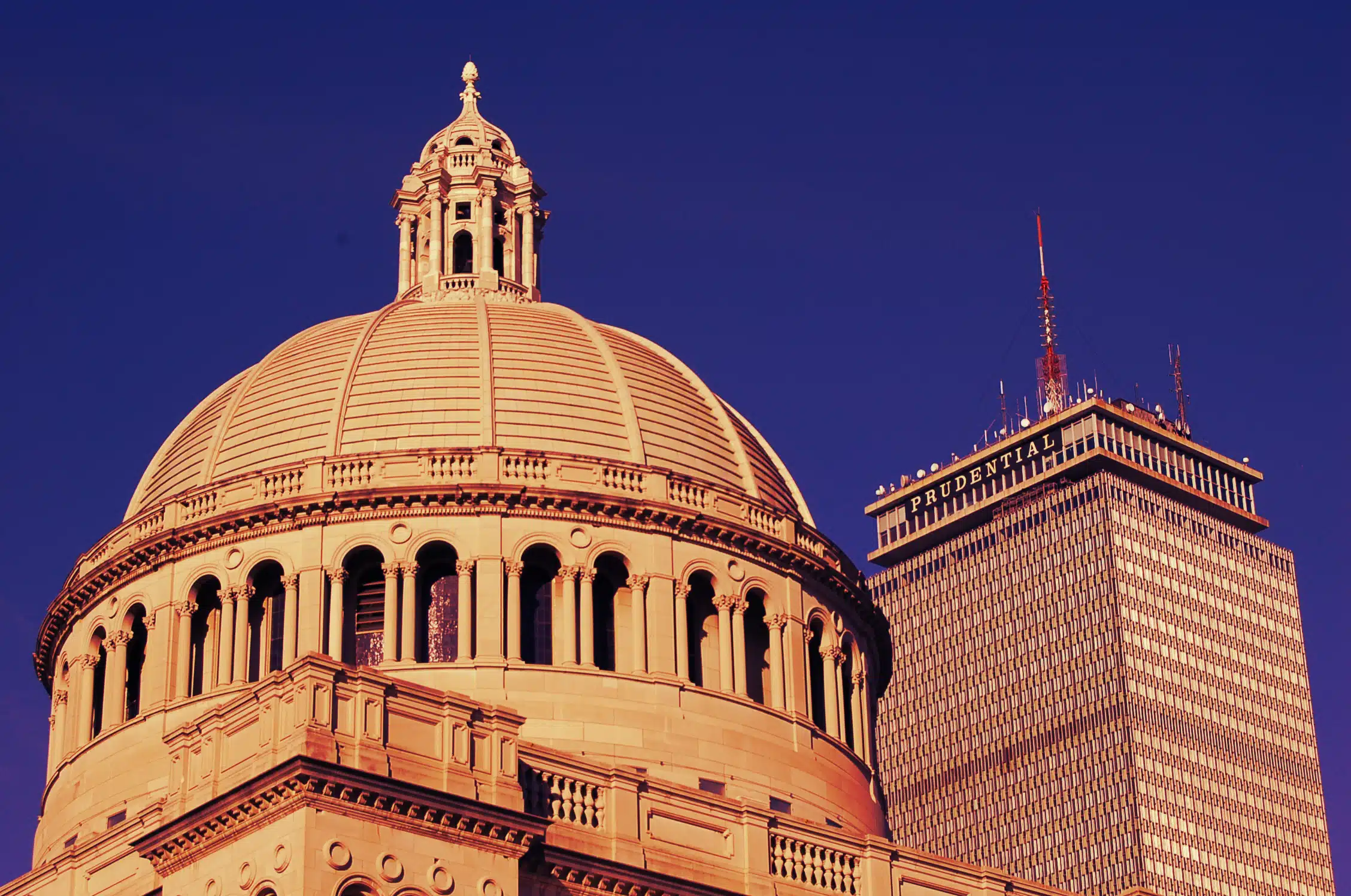 A domed building and a high-rise tower stand against a clear blue sky, basking in golden sunlight.