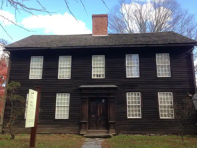 A historic two-story wooden house with symmetrical windows, a central entrance, and a prominent chimney. A sign is visible on the left side of the house. Trees surround the area.