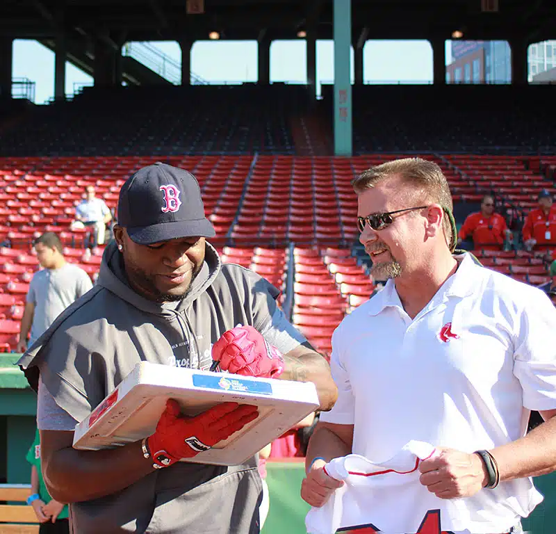 Two men are standing together in a baseball stadium. The man on the left, wearing a Boston Red Sox cap and gloves, is signing a base. The man on the right, in a white shirt, is holding a baseball jersey.