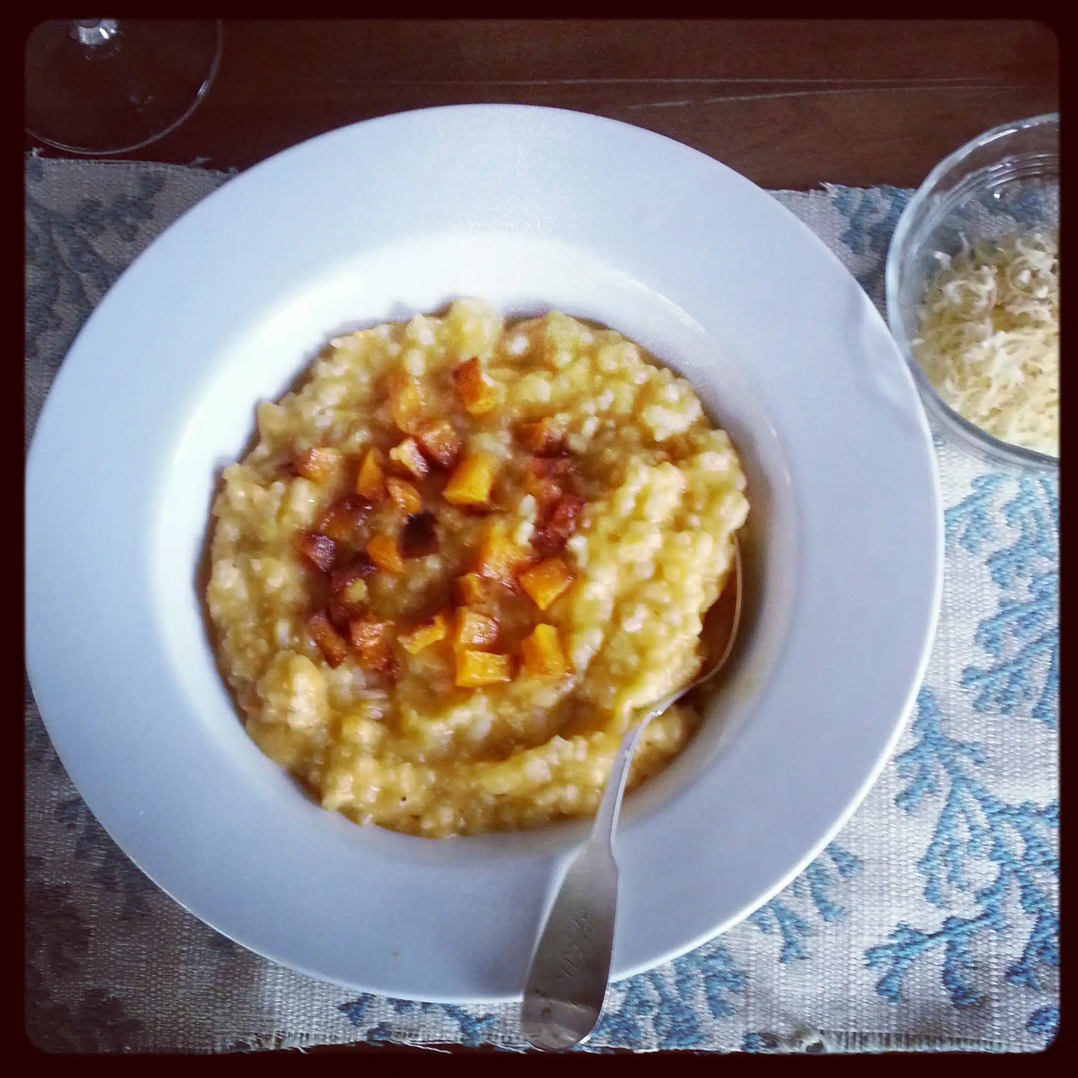 A bowl of risotto topped with diced pumpkin on a white plate, placed on a blue and white patterned tablecloth. A small bowl of grated cheese is placed nearby.