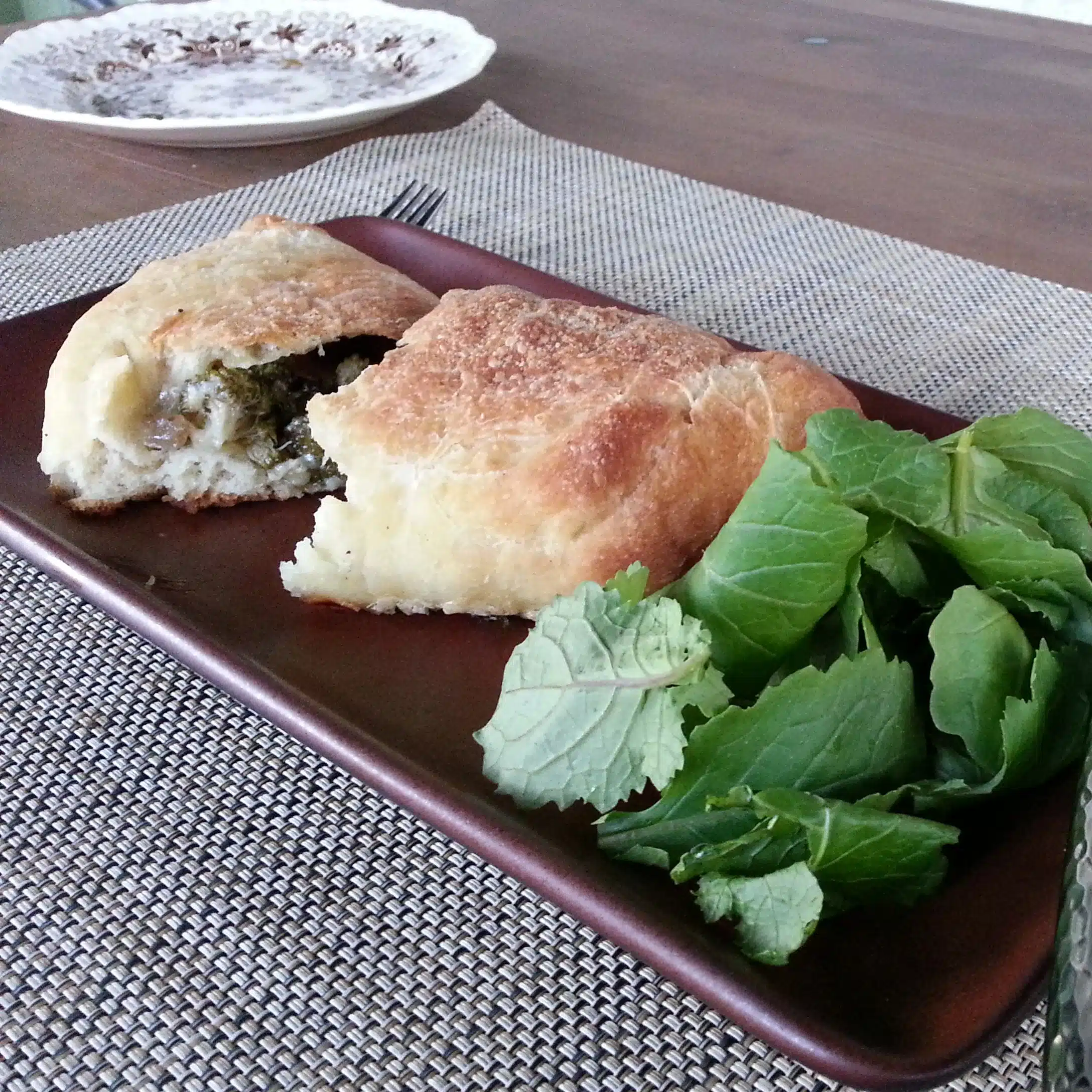 A partially sliced stuffed bread roll with a side of leafy greens on a brown rectangular plate. A fork and a round dish are also visible on a woven placemat.