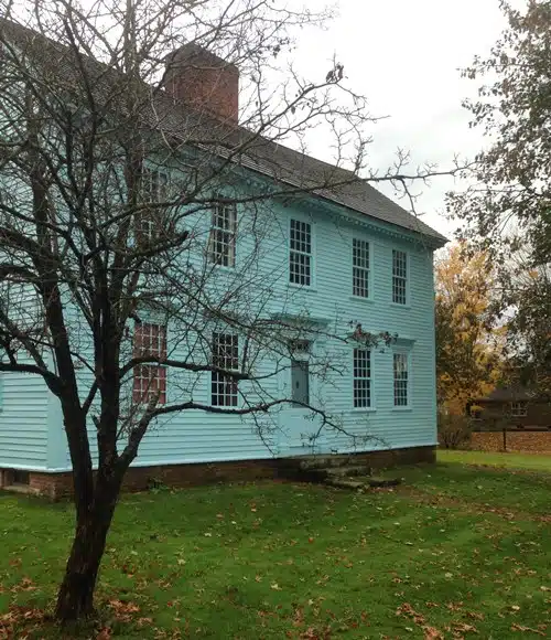 A light blue, two-story wooden house with numerous windows stands beside a leafless tree on a grassy lawn.
