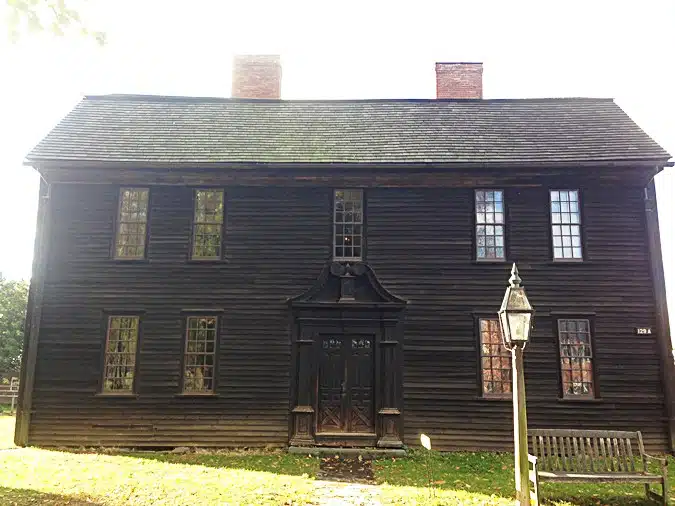 A large, dark wooden house with two chimneys and multiple windows, featuring a central front door with ornate details. A lamp post and bench are in the front yard.