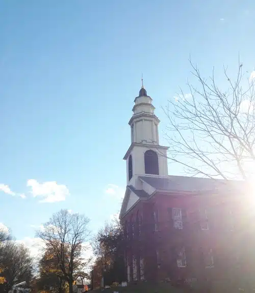 A brick building with a white steeple and clock tower, partially obscured by sunlight, stands against a clear blue sky with a few scattered clouds and trees without leaves in the foreground.