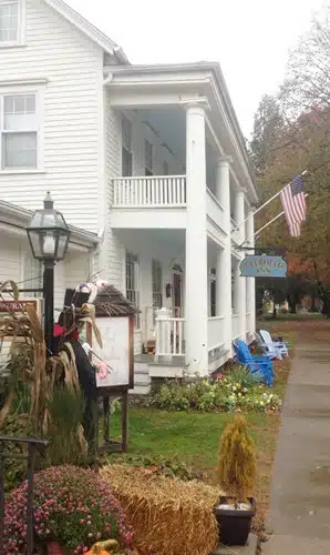 White two-story building with colonial pillars and an American flag. A “Cracker Barrel” sign hangs near the entrance. A porch with blue chairs, a lamp post, and a seasonal scarecrow are visible.
