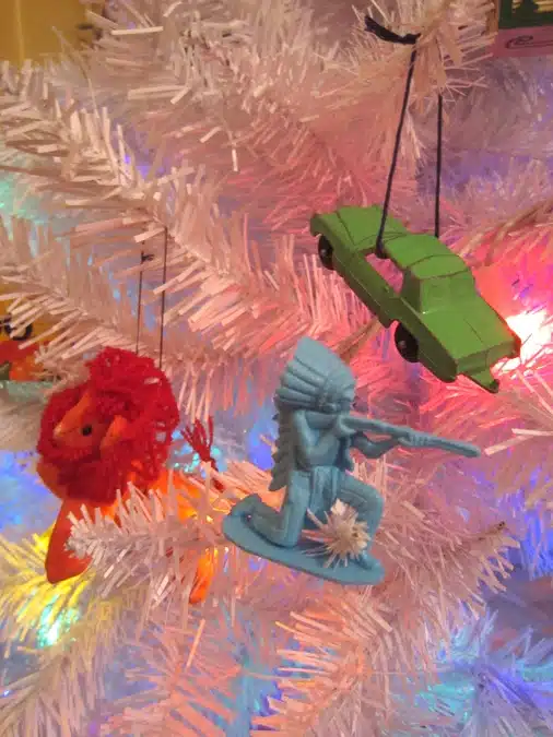 Ornaments on a white Christmas tree include a lion figure, a blue toy soldier aiming a gun, and a green car.