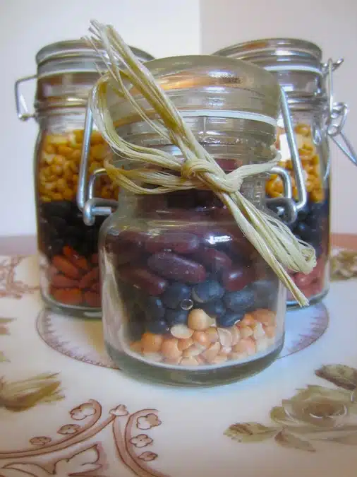 Three glass jars filled with various dried beans and lentils, each sealed with a metal clasp, and one jar adorned with a tied straw ribbon in front. All jars are placed on a decorative surface.
