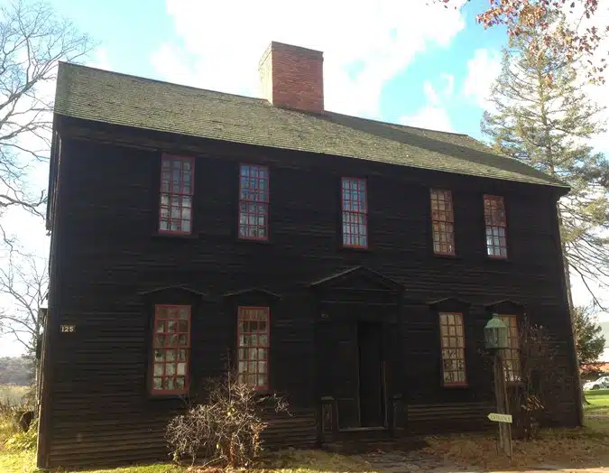 A large, dark-colored, two-story historical house with a brick chimney, red-framed windows, and a central front entrance. Sparse foliage surrounds the building.