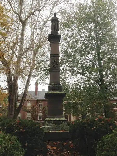 A tall stone monument with a statue on top stands in a park area surrounded by trees and buildings in the background.