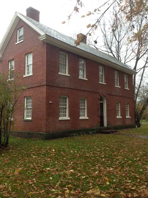 A two-story red brick house with a white window trim, surrounded by autumn leaves on the ground and bare trees in the background.