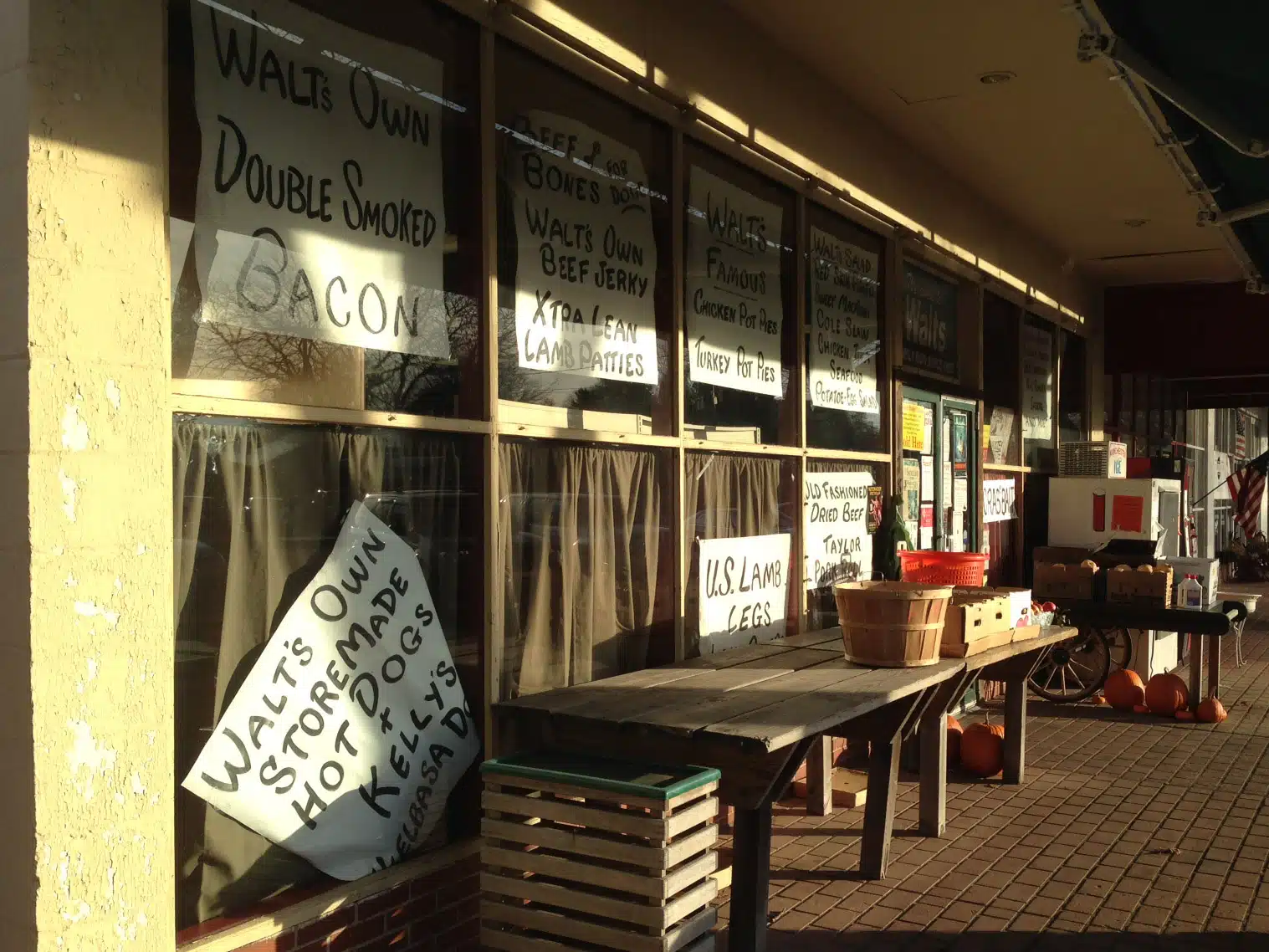 A storefront with various hand-written signs advertising products like bacon, beef jerky, and hot cocoa. Tables outside hold baskets and pumpkins, while an American flag is partially visible on the right.