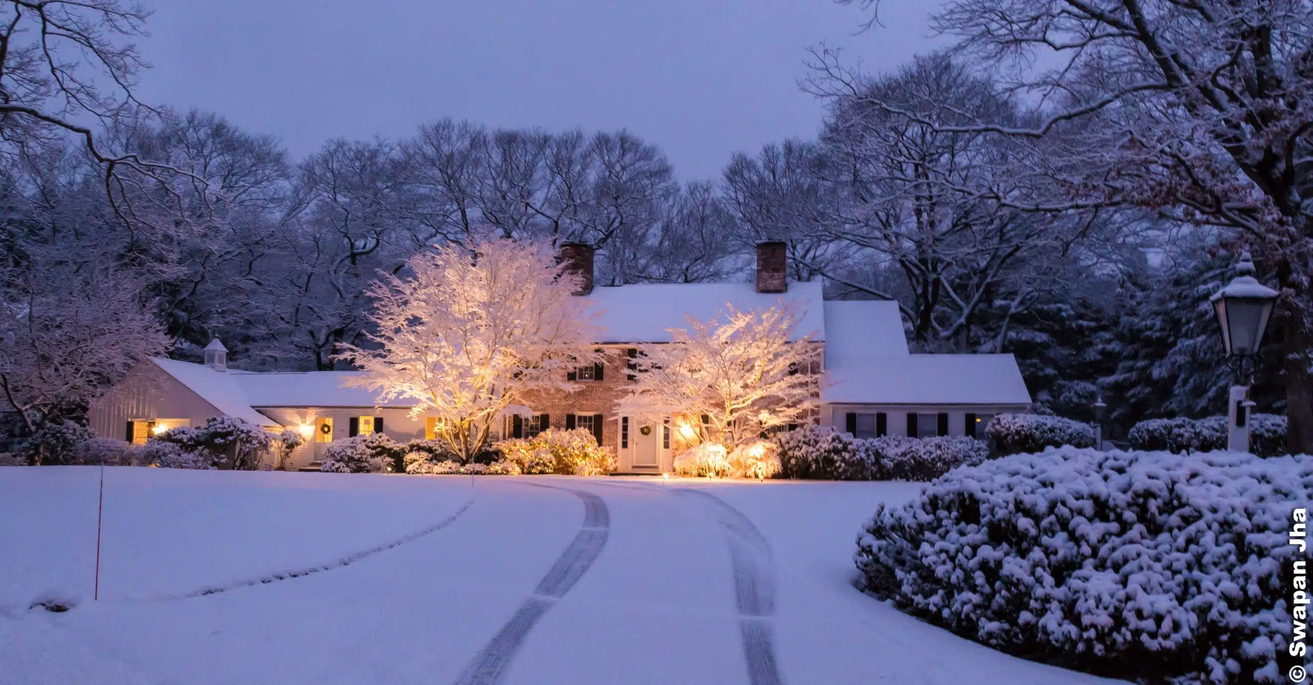 A snow-covered house with illuminated trees in front, surrounded by a snowy landscape, featuring a driveway with tracks leading up to the entrance.