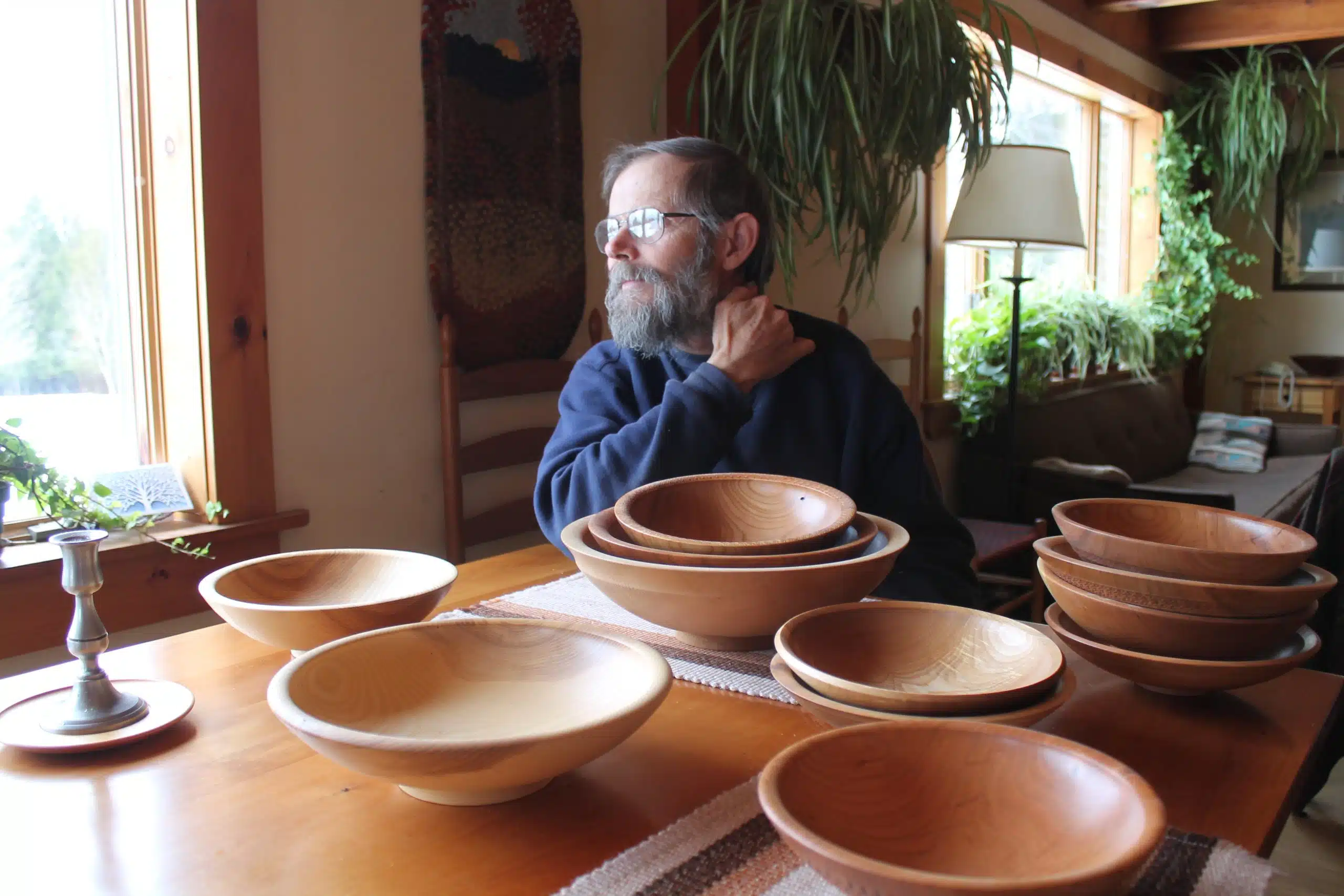 A person with a beard and glasses sits at a wooden table inside a room surrounded by several wooden bowls of different sizes, with plants and a large window in the background.