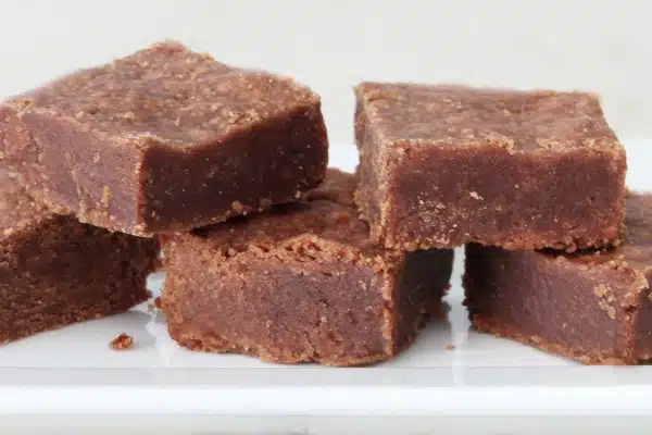 A stack of six chocolate brownies is displayed on a white plate. The brownies have a dense and moist texture with a slightly crumbly top.