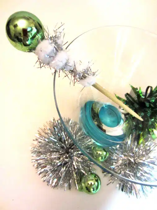 A clear glass bowl contains a blue liquid and is decorated with silver tinsel, green ornaments, and a festive stir stick with green, white, and silver elements. Silver and green decorations are beside it.