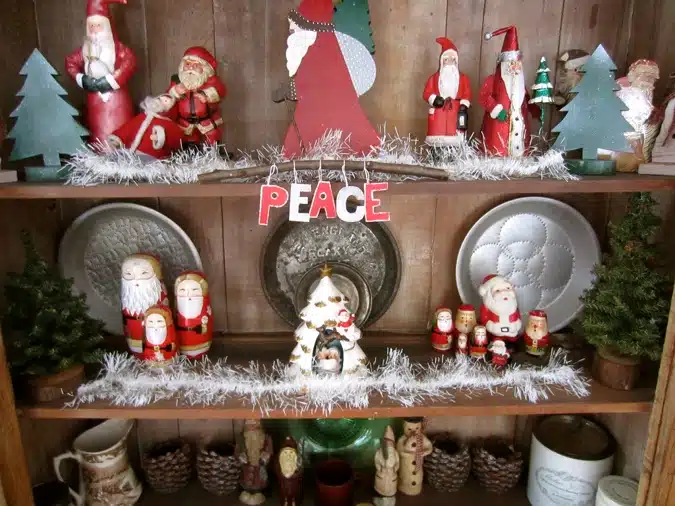 Wooden shelves adorned with various Santa Claus figurines, two small Christmas trees, metal plates, and a sign with the word "PEACE" hanging from white garland.