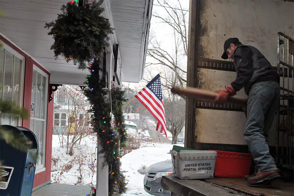 A postal worker stands in a mail truck loading a package, with an American flag and Christmas decorations visible. Snow is on the ground, suggesting a winter setting.