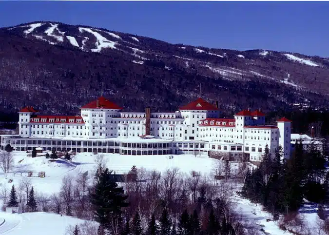 Large white hotel with red roofs surrounded by snow-covered landscape and forested mountains in the background.