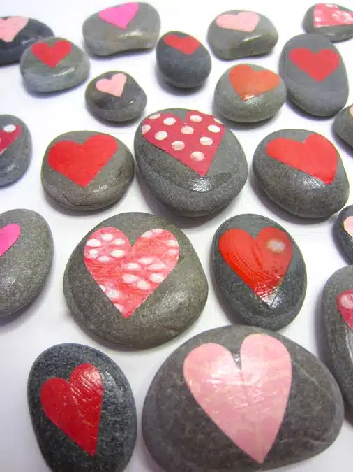 Several gray stones are scattered on a white surface, each adorned with painted red or pink hearts, some solid and others with patterns.