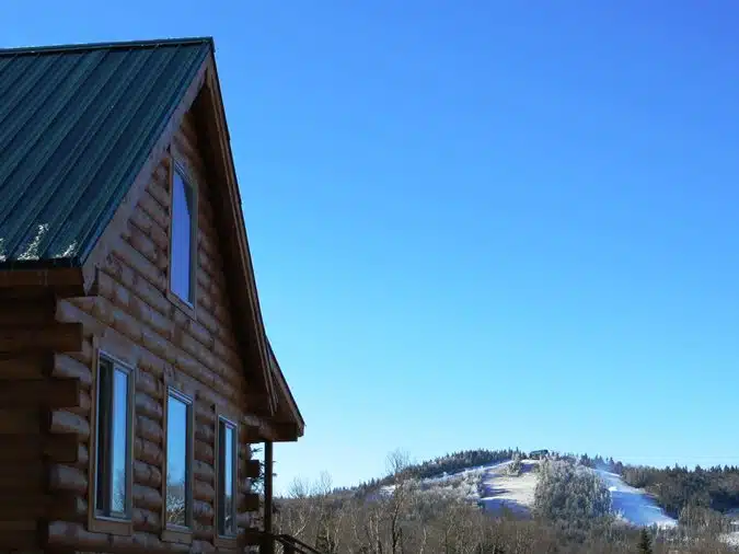 A wooden cabin with a green roof on the left, with a snow-covered hill and clear blue sky in the background.