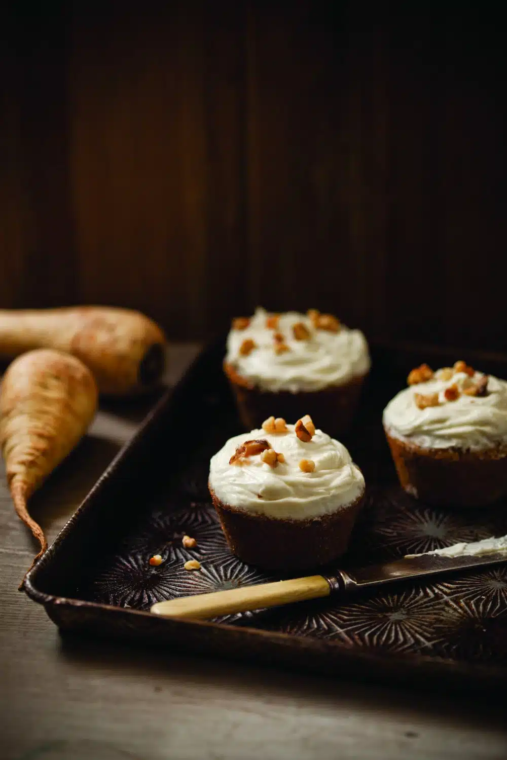Three frosted cupcakes topped with nuts on a dark tray, accompanied by a knife and a couple of parsnips in the background.