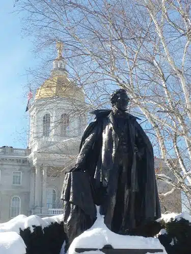 A bronze statue stands in front of a building with a golden dome on a clear winter day, surrounded by leafless trees and snow.