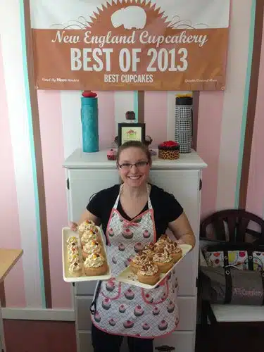A person holding two trays of cupcakes, standing in front of a sign that reads "New England Cupcakery Best of 2013." The room has pink and white striped walls and a cabinet with decorative items.