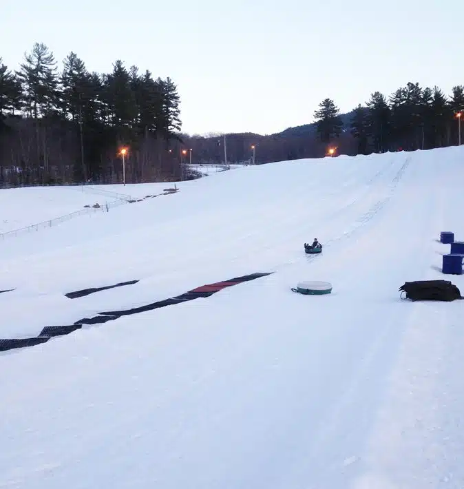 A snow-covered hill with multiple tubing lanes, surrounded by evergreen trees, features a person tubing down one lane. The area is illuminated by overhead lights.