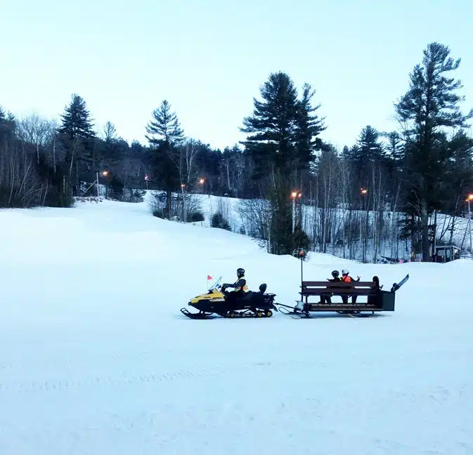 A snowmobile pulling people seated on a wooden sleigh across a snow-covered landscape surrounded by trees.