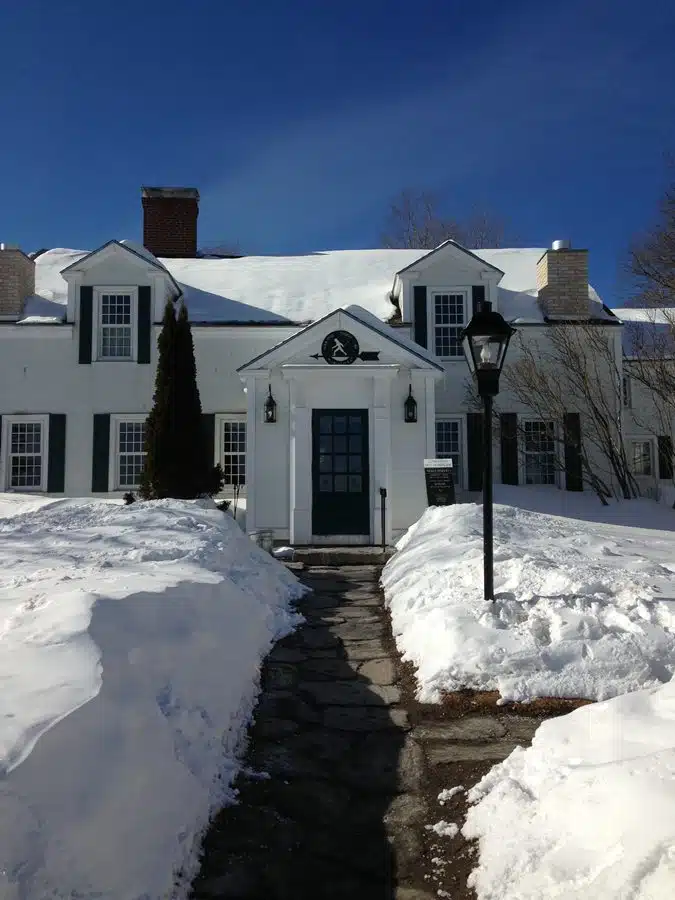 White house with snow-covered roof and lawn, dark blue shutters, small trees, and a stone path leading to a dark blue front door. A black lamp post stands near the right side of the path.