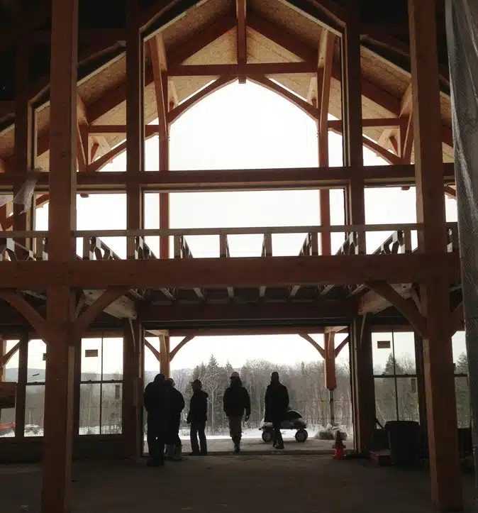 Interior of a large wooden building under construction with a high vaulted ceiling; five silhouetted people stand near the entrance, looking outside.