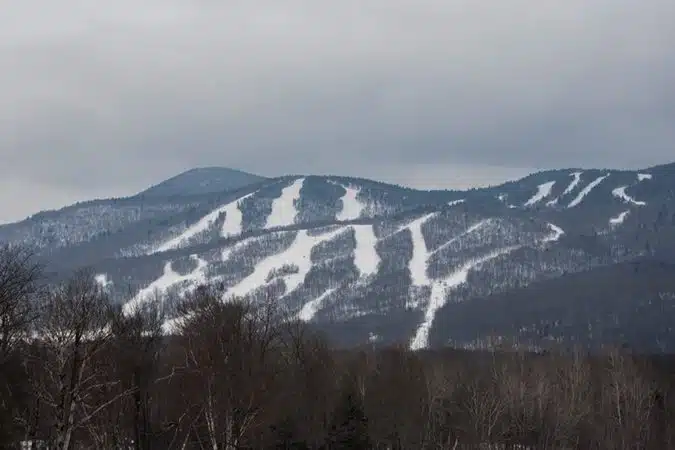 Snow-covered ski trails are visible on a mountain side, surrounded by leafless trees under a cloudy sky.