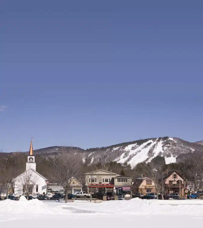 A small town with a white church and several buildings is in front of a snow-covered mountain and ski slope under a clear blue sky.