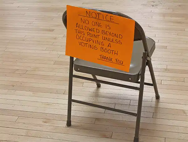 A folding chair on a wooden floor has an orange sign attached, stating, "NOTICE: No one is allowed beyond this point unless occupying a voting booth. Thank you.